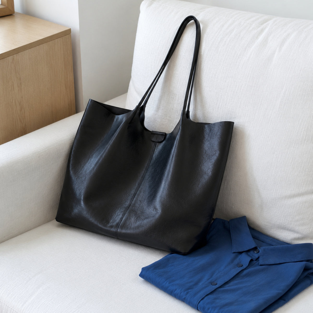 Black Vegan PU Leather tote resting on a white sofa beside a folded blue shirt