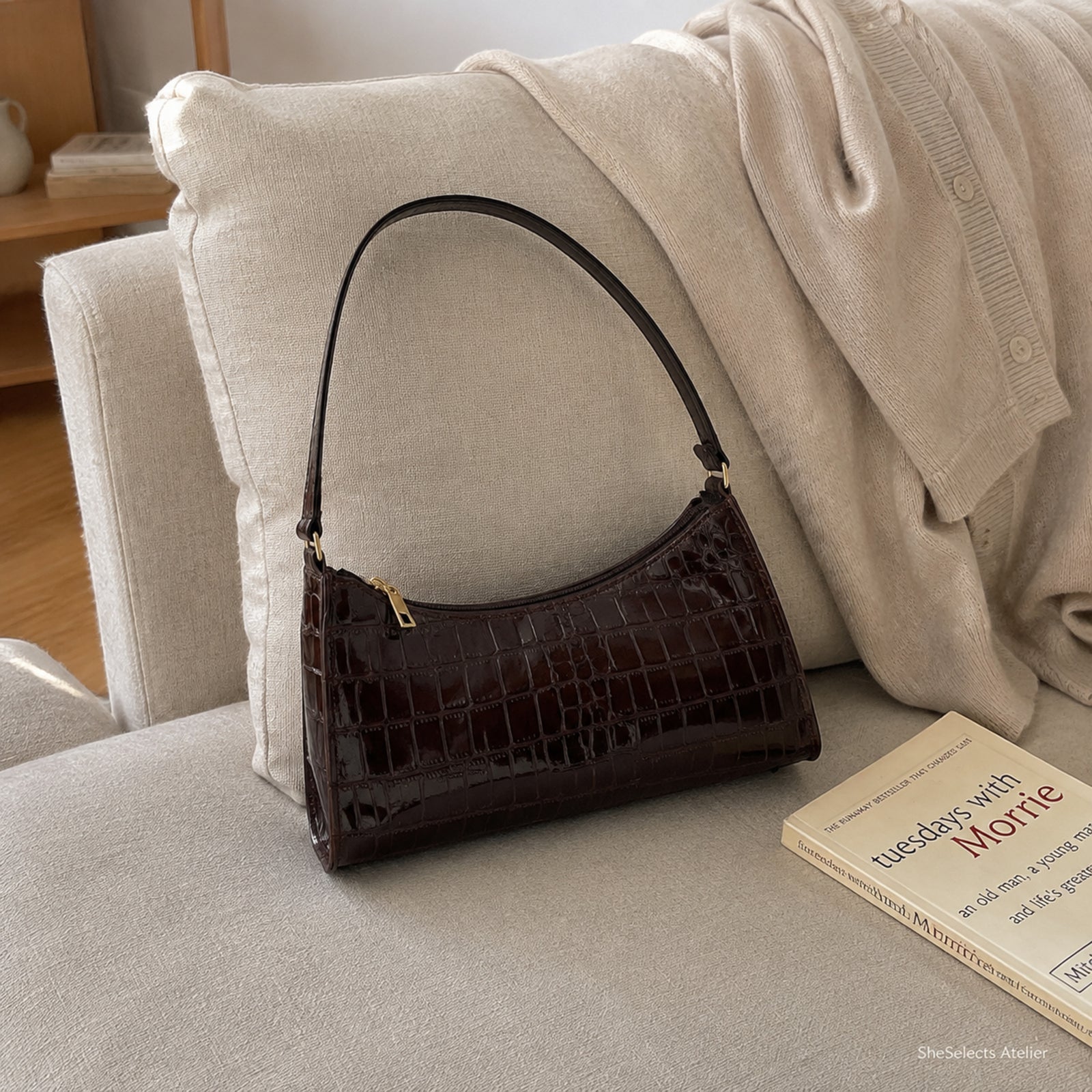 Dark brown glossy mini shoulder bag on a beige linen sofa beside a cardigan and book