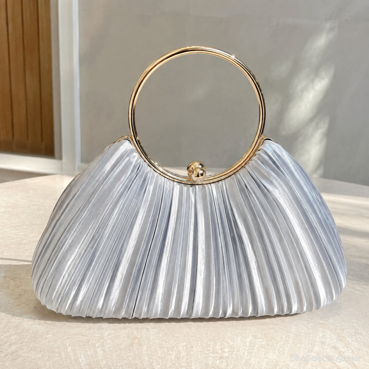 Silver pleated satin evening clutch with circular gold ring handle in natural window light
